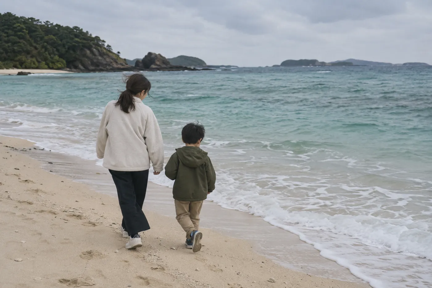 冬の曇りの日の南の島の砂浜を、上着を着た母親と男の子が歩いているイメージ